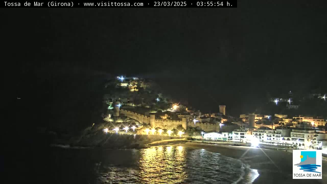 A nighttime coastal town with a walled castle on a hill overlooking a beach and calm water.
