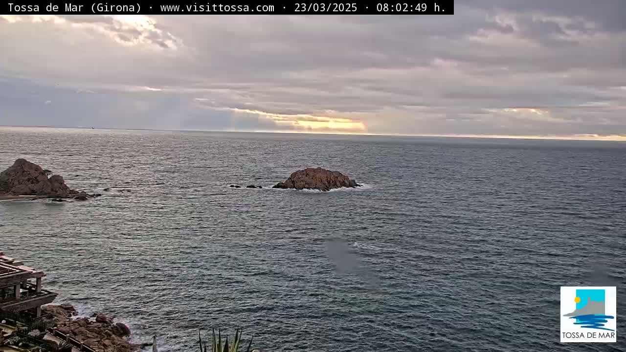 A rocky outcrop and a distant coastline are visible on a partly cloudy day over a dark, calm sea.