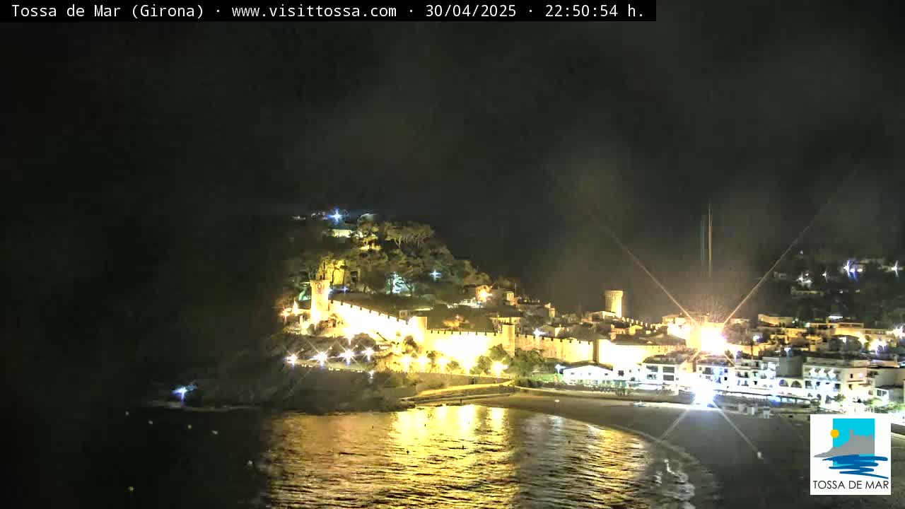 A nighttime view of a coastal town with a walled castle, illuminated buildings, and a calm, reflective body of water.