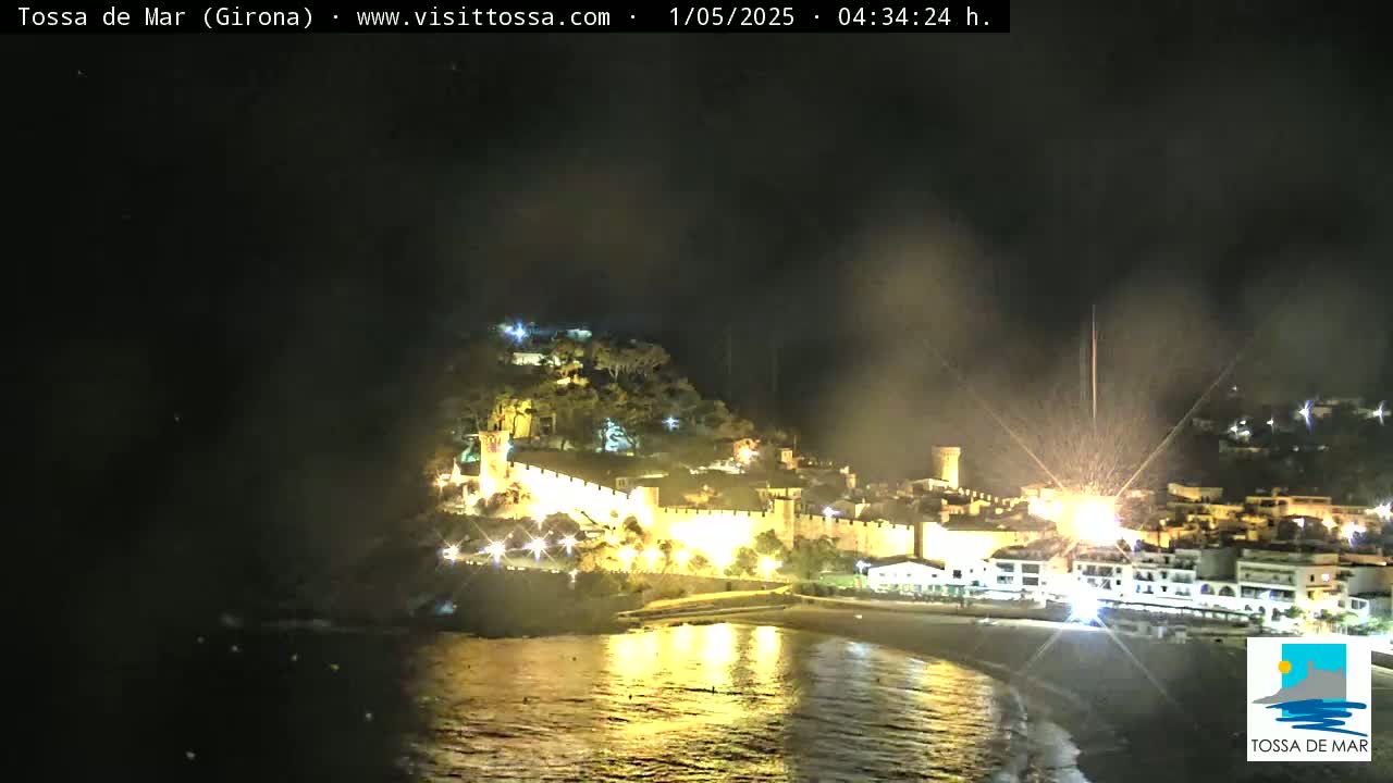 A nighttime view of a coastal town with a fortress illuminated along the water's edge under a dark sky.