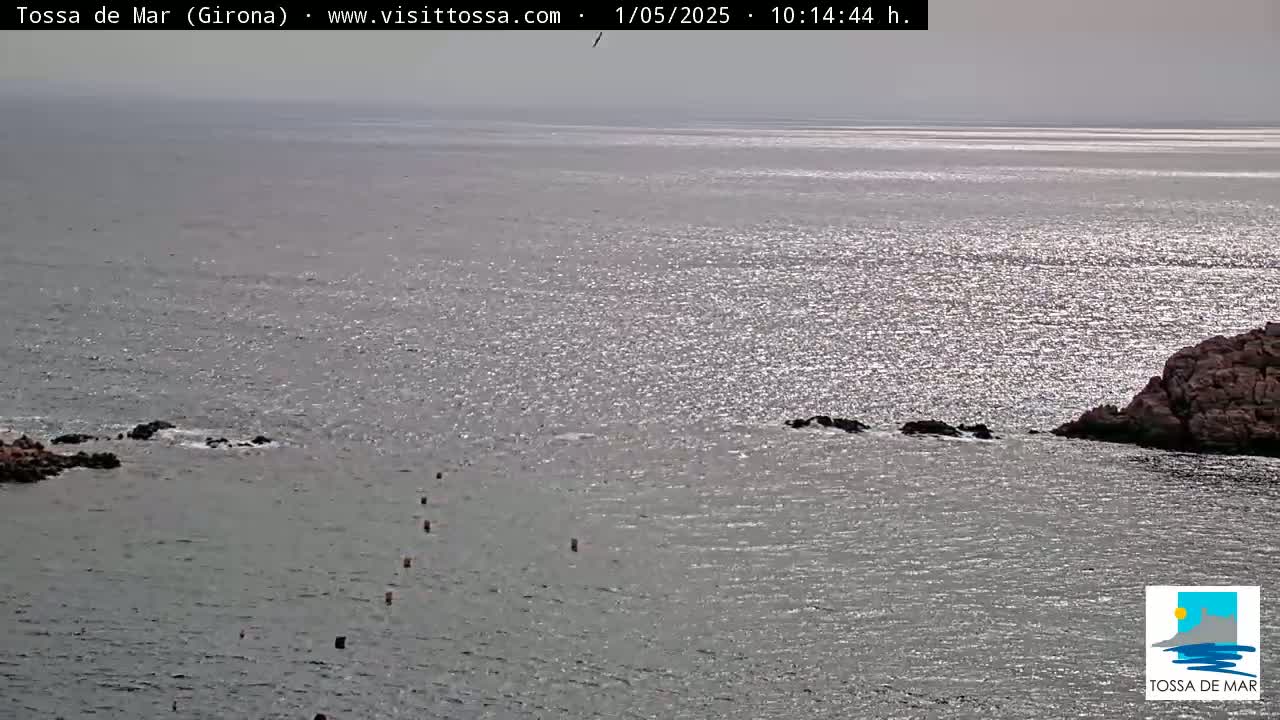 A slightly hazy, sunny day shows a calm sea with sparkling water, rocky outcrops, and a few buoys in the water.