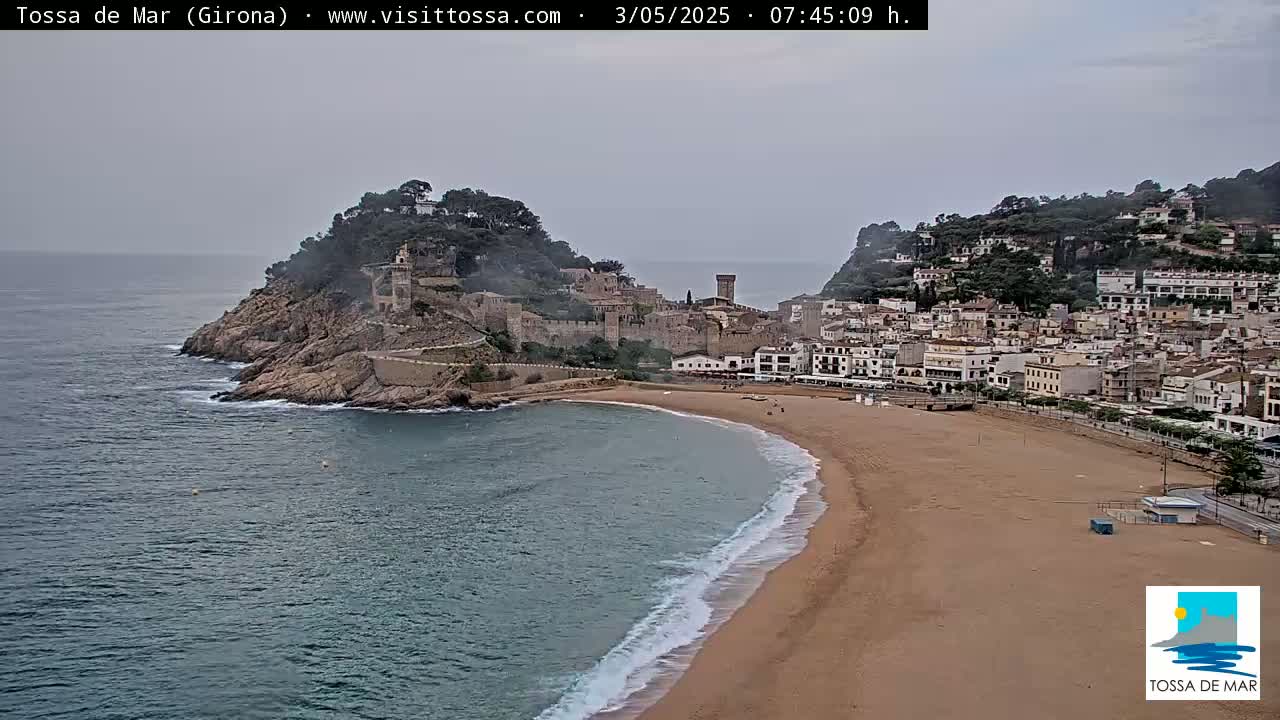 A coastal town with a sandy beach and medieval-looking buildings nestled on a cliffside overlooking a calm sea under an overcast sky.
