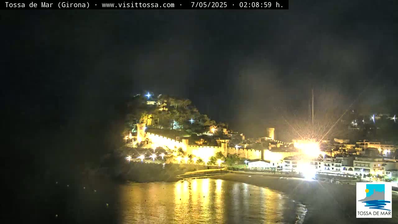 A nighttime coastal town with a walled fortress is illuminated, reflecting in the dark water under a clear sky.