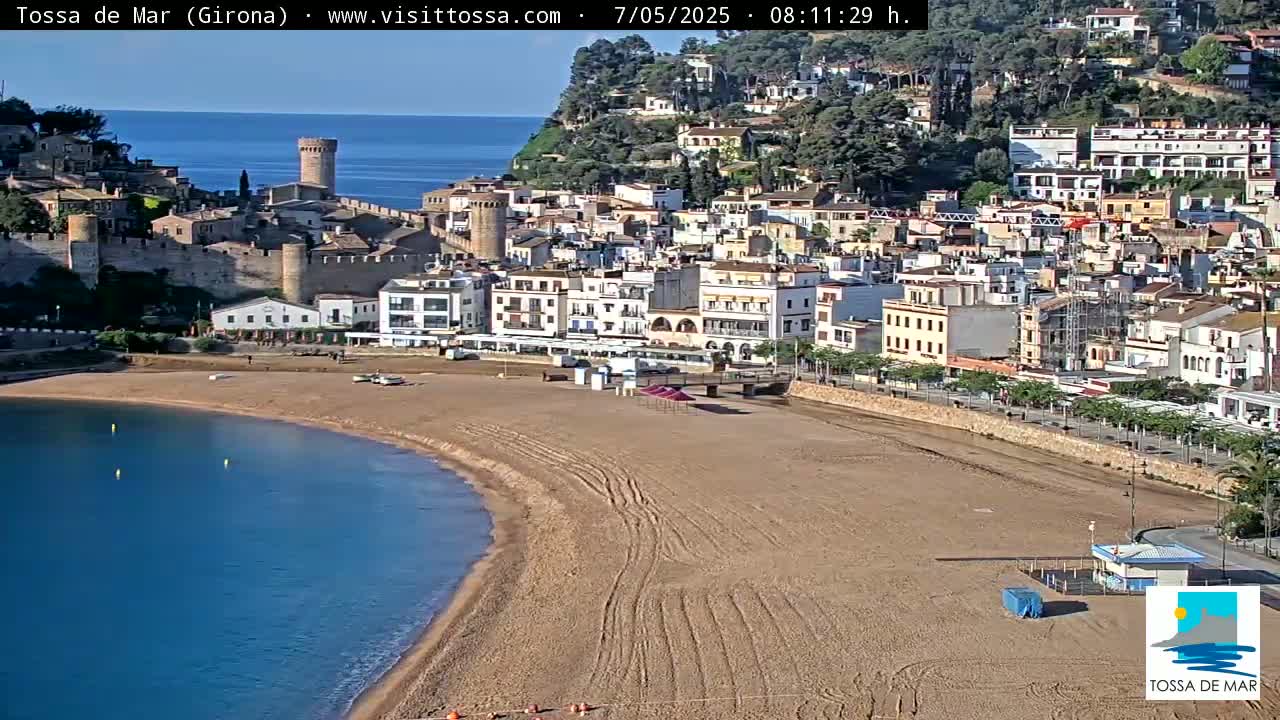 A sandy beach curves into a calm blue bay, bordered by a town with medieval walls and multi-story buildings climbing a hillside under a clear sky.