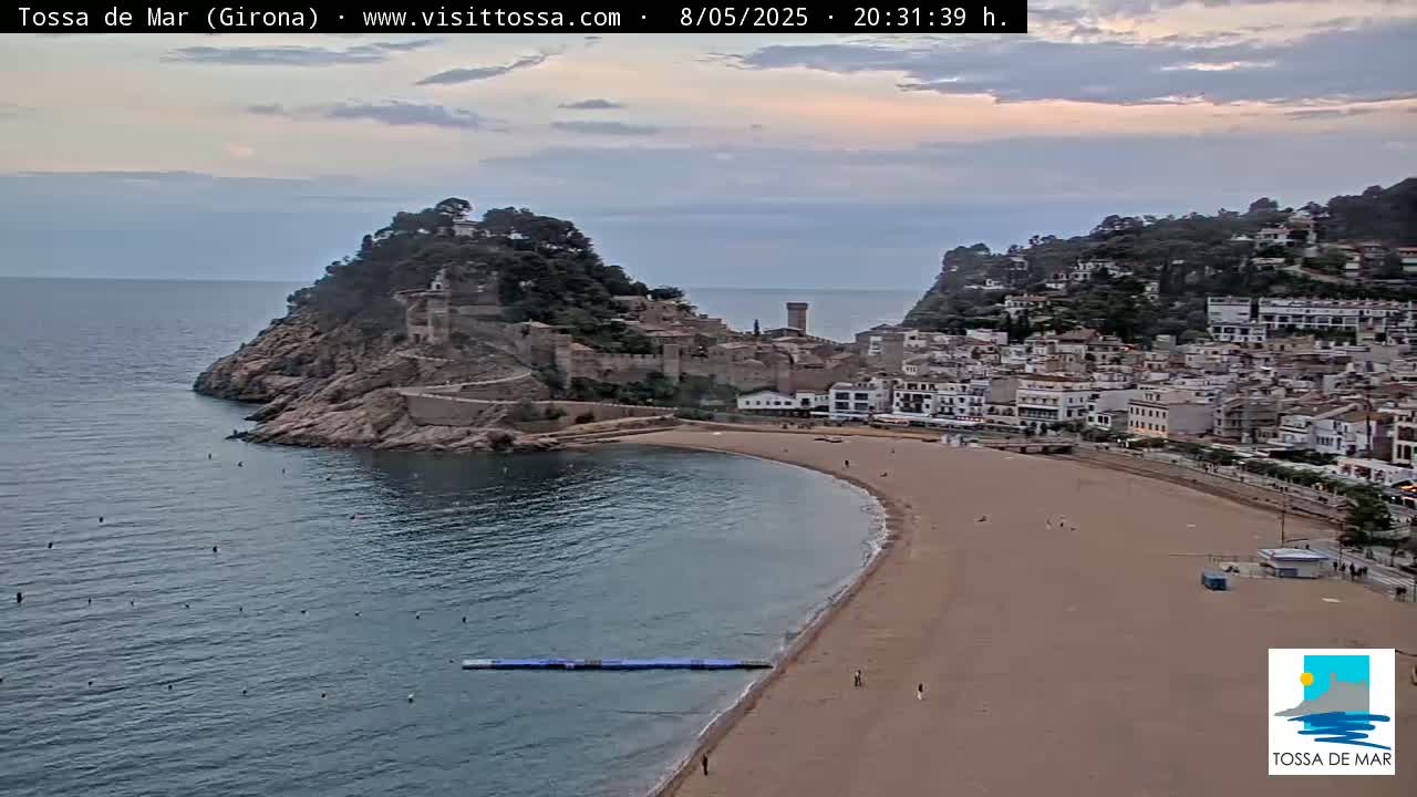 A sandy beach curves around a calm bay, with a medieval-style town perched on a cliff overlooking the water under a partly cloudy, dusky sky.