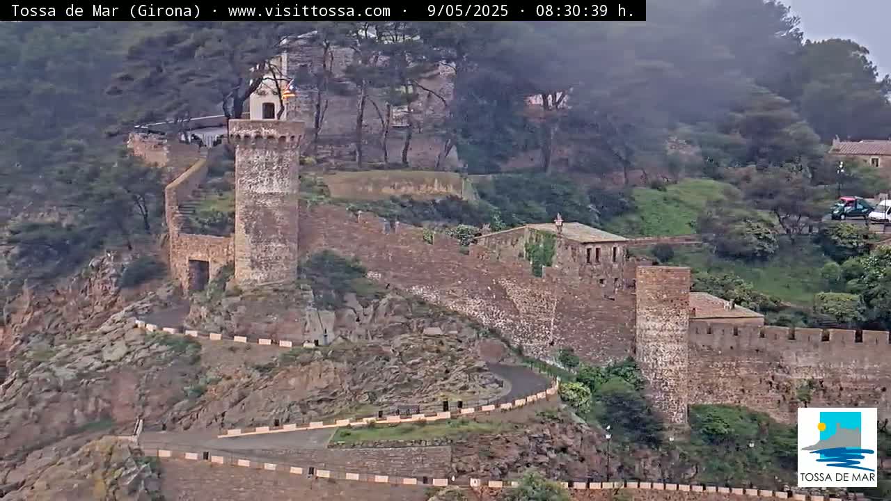 A stone tower and wall are perched on a rocky hillside overlooking a winding road, under an overcast sky.