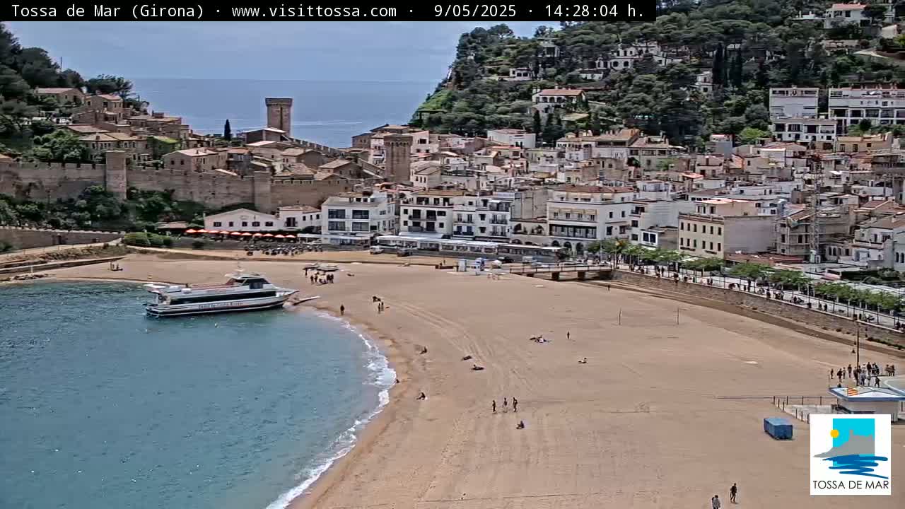 A sandy beach curves along a clear blue body of water in front of a coastal town nestled against a hillside on a sunny day.