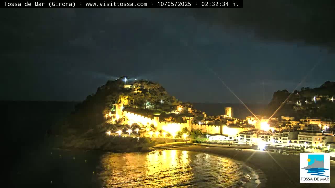 A nighttime coastal town, illuminated, sits on a hill overlooking a dark ocean under a mostly clear sky.