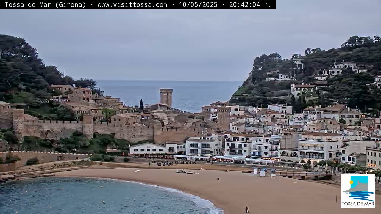 A coastal town with a sandy beach and medieval-style buildings sits on a hill overlooking a calm sea under an overcast sky.