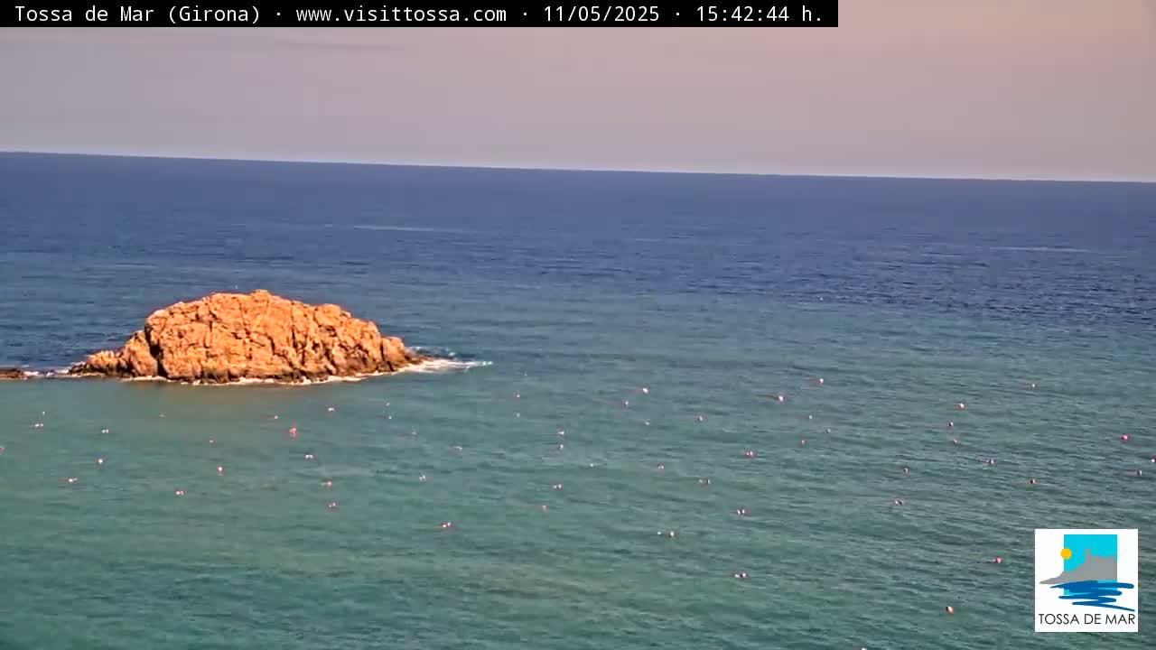 A rocky outcrop sits in calm, clear teal water dotted with small, pink buoys under a pale blue sky.