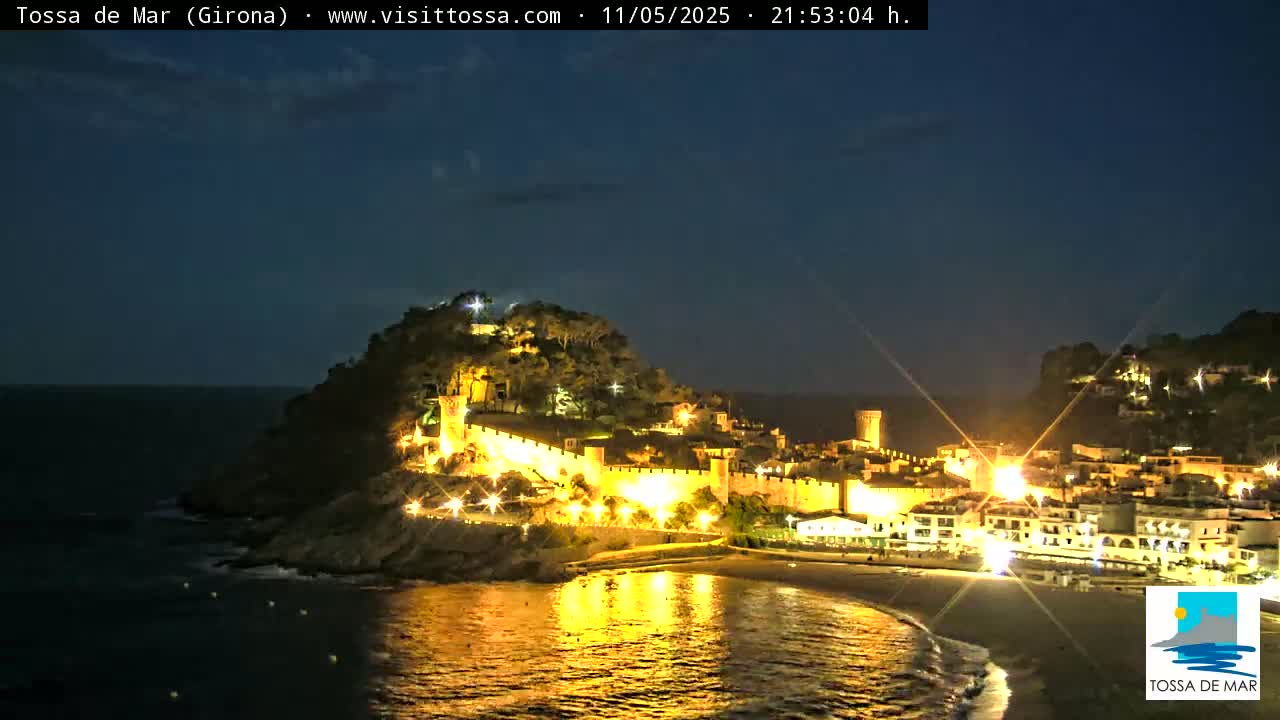 A coastal town, illuminated at night, sits nestled on a hill overlooking a calm, dark ocean under a clear, twilight sky.