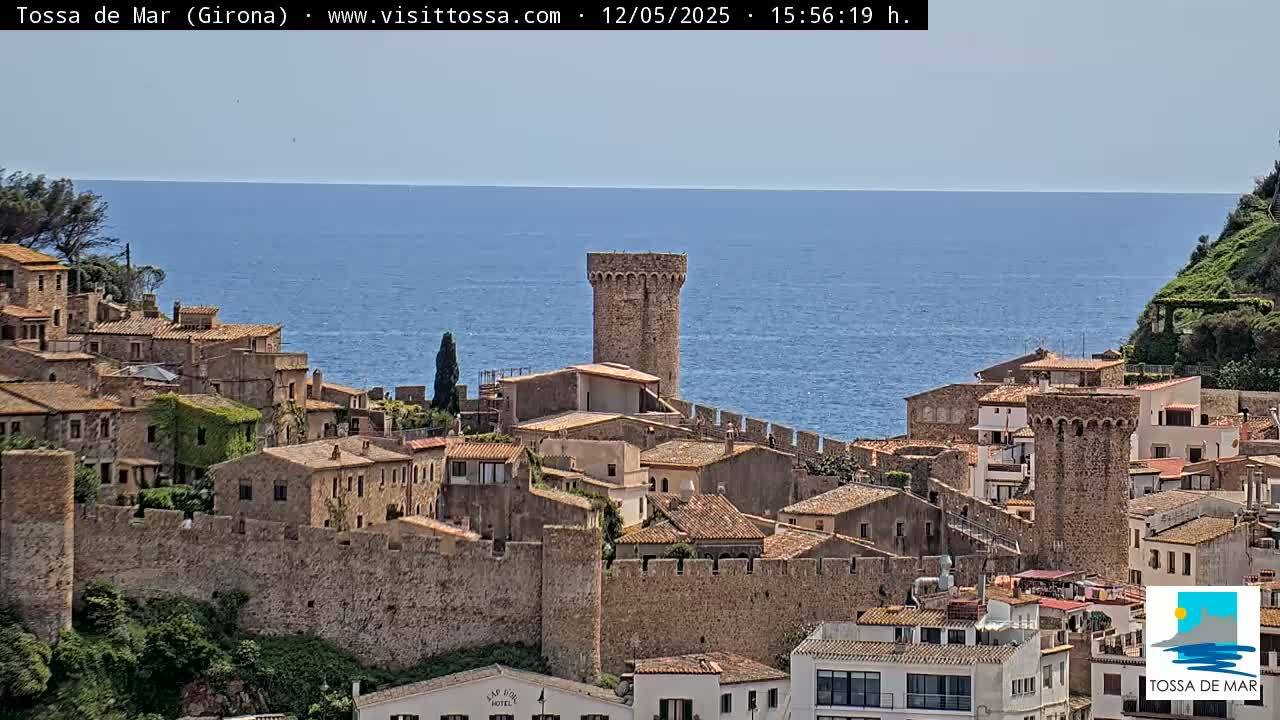 A coastal town with stone buildings and walls overlooks a calm blue sea under a clear sky.
