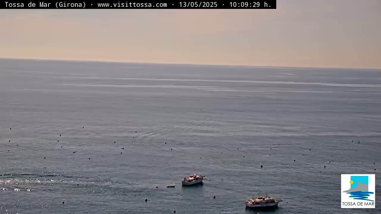 A calm, light-grey sea is speckled with small, dark objects, and two small boats are visible in the lower center.