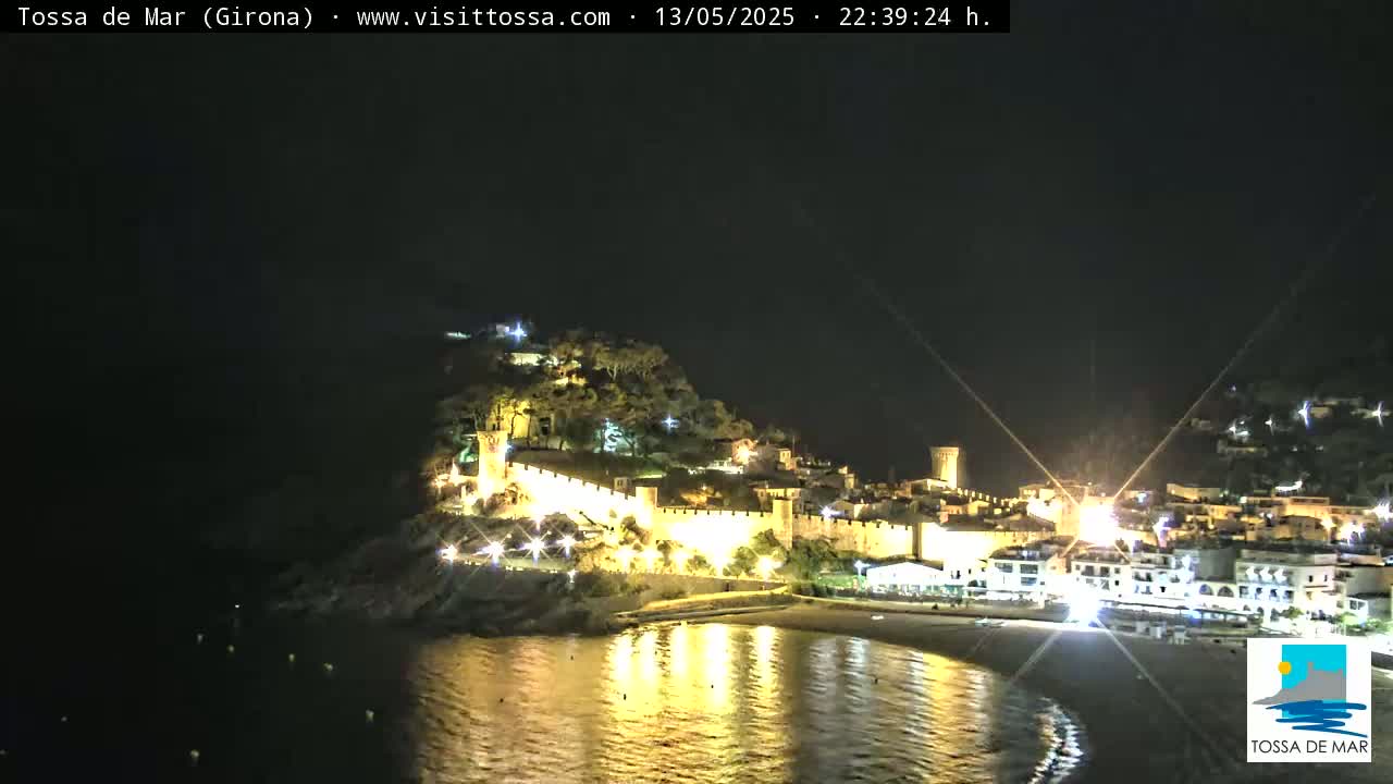 A nighttime view of a coastal town with a walled fortress, illuminated buildings, and calm water reflecting the lights.
