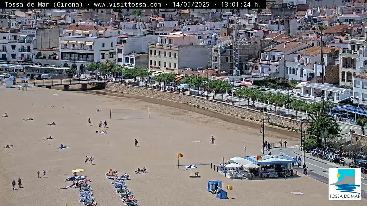 A sandy beach with a few people scattered across it, in front of a town with white buildings under a bright sunny sky.