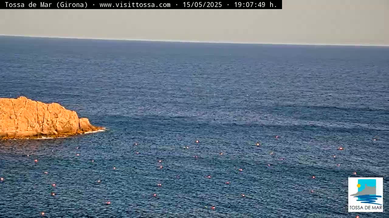 A rocky outcrop juts out into a calm, blue sea dotted with small, orange buoys under a mostly clear sky.