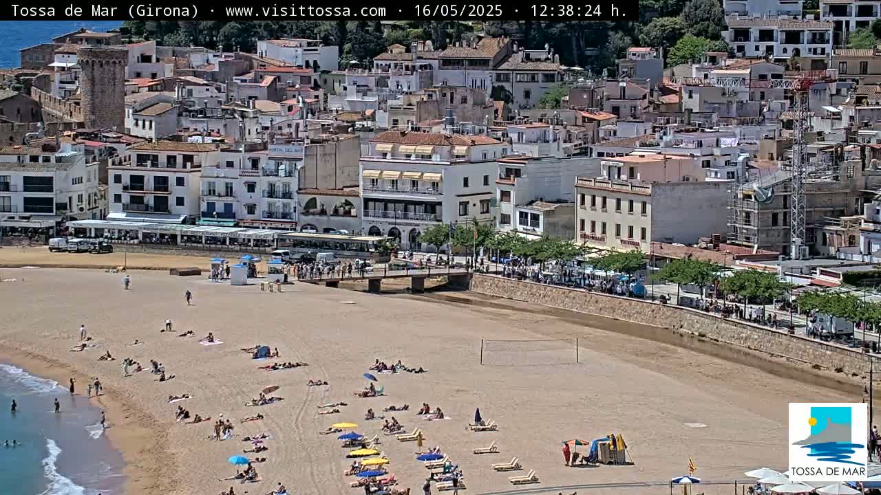 A sandy beach with many sunbathers and a small town with buildings and a stone tower are visible on a sunny day.