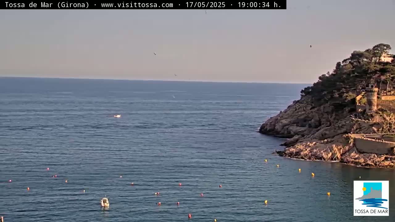 A calm, blue sea laps against a rocky shoreline under a clear, light-blue sky, with a few birds flying overhead and buoys scattered in the water.