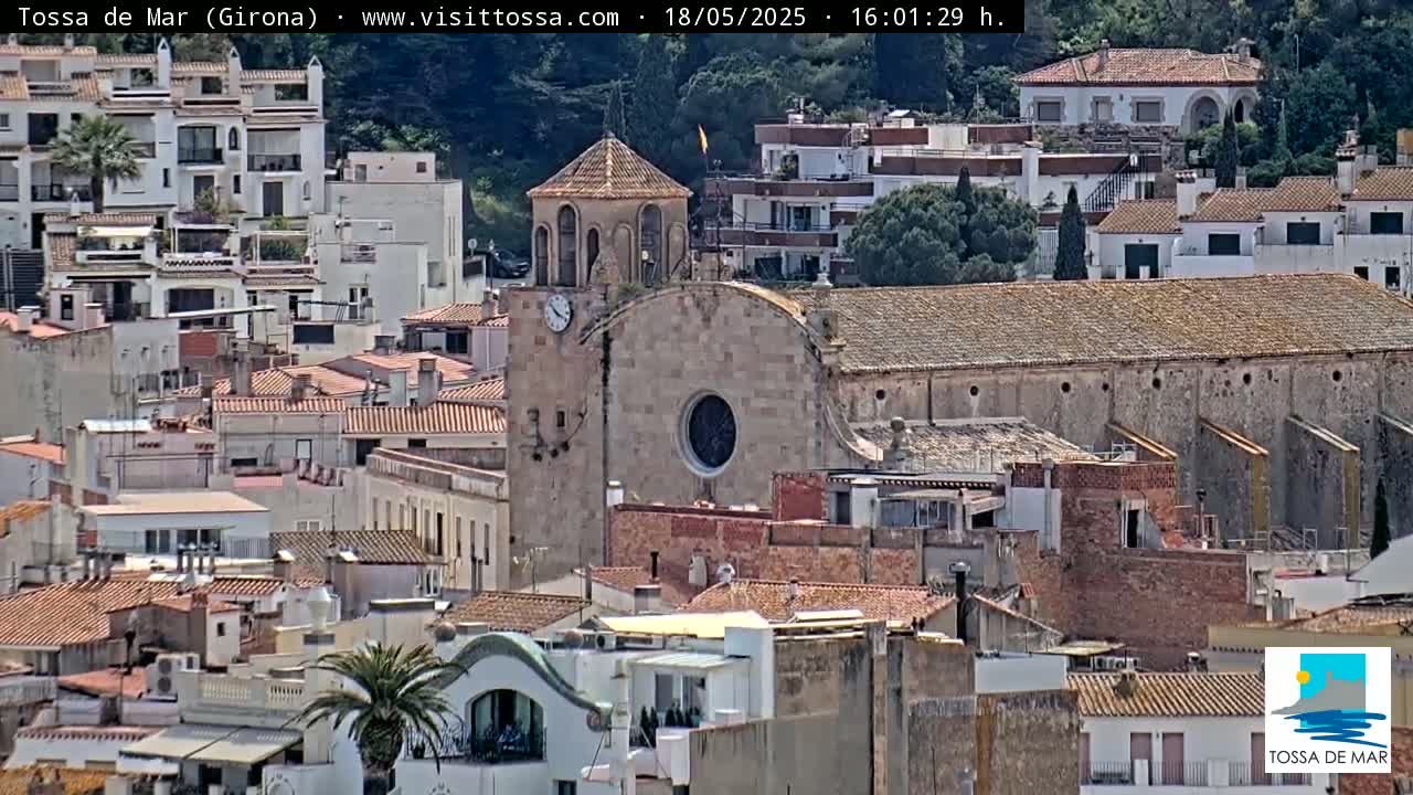 A church and many buildings with red-tiled roofs are clustered together on a hillside on a sunny day.