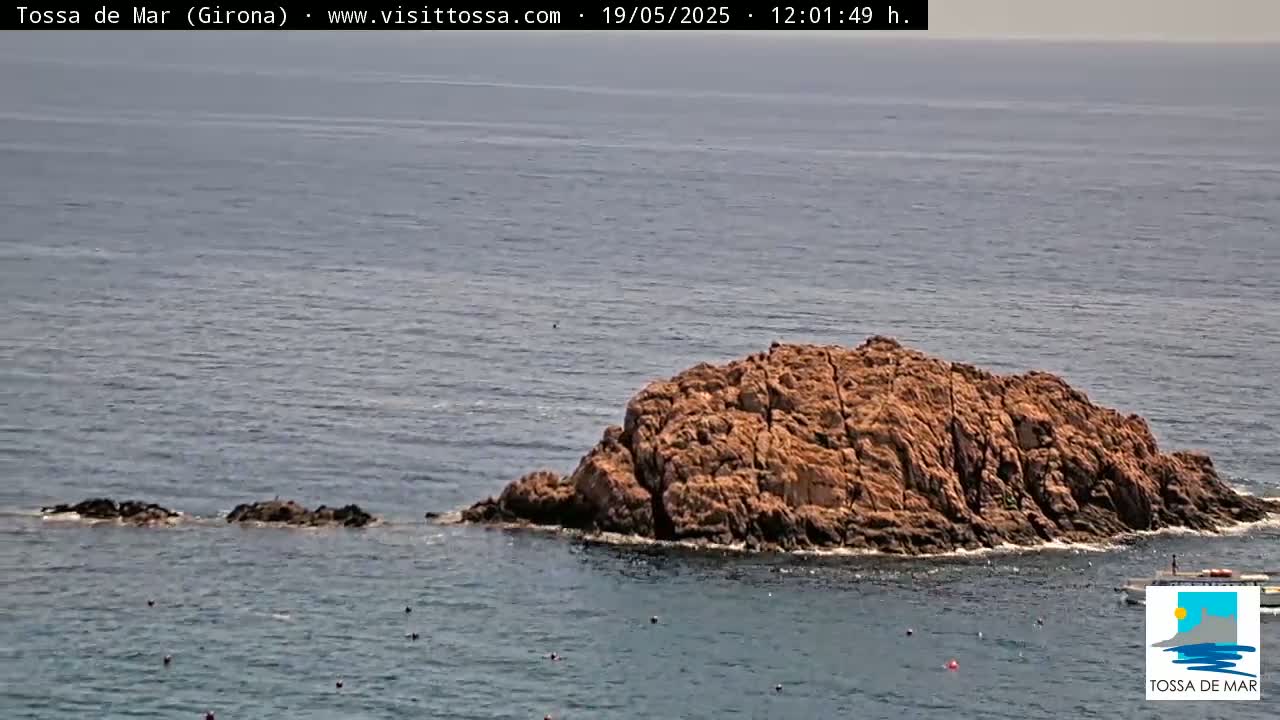 A rocky outcrop and several smaller rocks jut out from a calm, blue sea under a mostly clear sky.