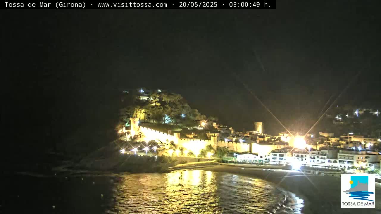A nighttime coastal town with a walled fortress is illuminated, reflected in calm water.