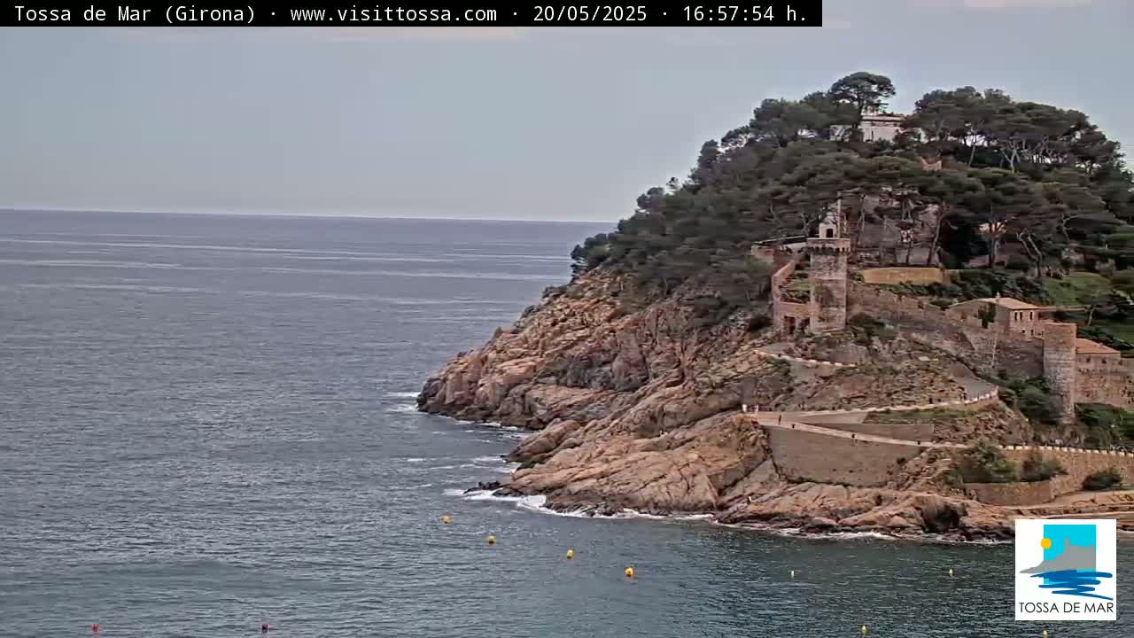 A rocky coastline with a medieval-style castle and surrounding buildings on a hill overlooking a calm sea under a mostly cloudy sky.