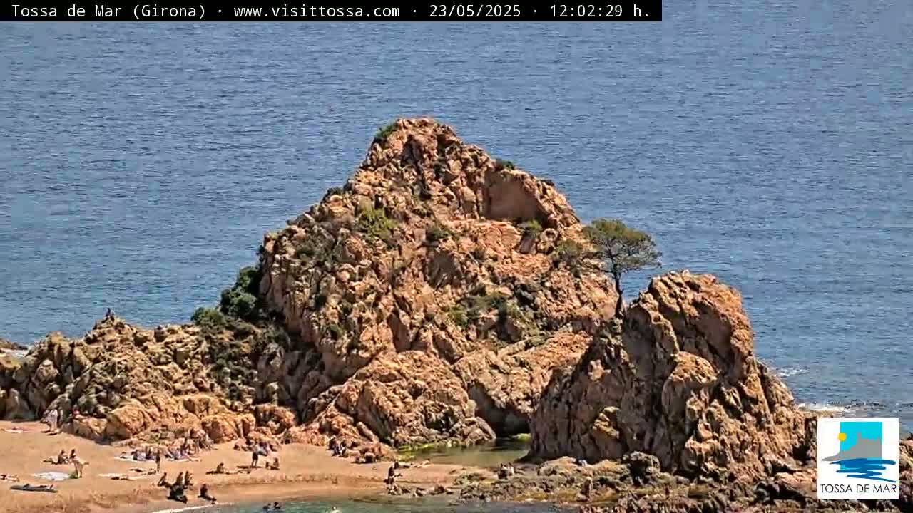 A rocky outcrop with a small sandy beach and scattered people sits on a calm, blue ocean under a sunny sky.