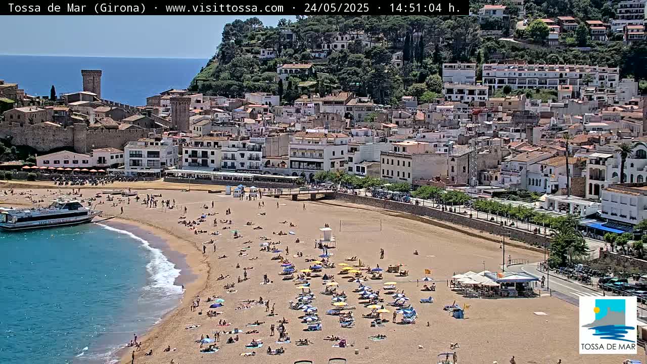 A sandy beach crowded with sunbathers is situated below a seaside town built on a hill, under a clear sunny sky.