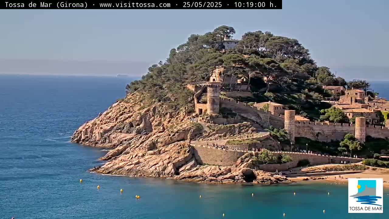 A coastal town with a medieval castle sits atop a rocky cliff overlooking a calm, blue sea on a sunny day.