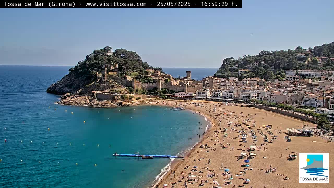 A sandy beach crowded with people curves around a calm, clear blue bay, next to a hilltop medieval castle under a bright, sunny sky.