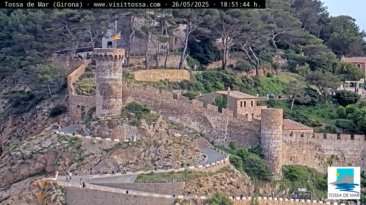 A stone wall and towers, part of a castle or fortress, winds along a hillside covered in green vegetation under a partly cloudy sky.