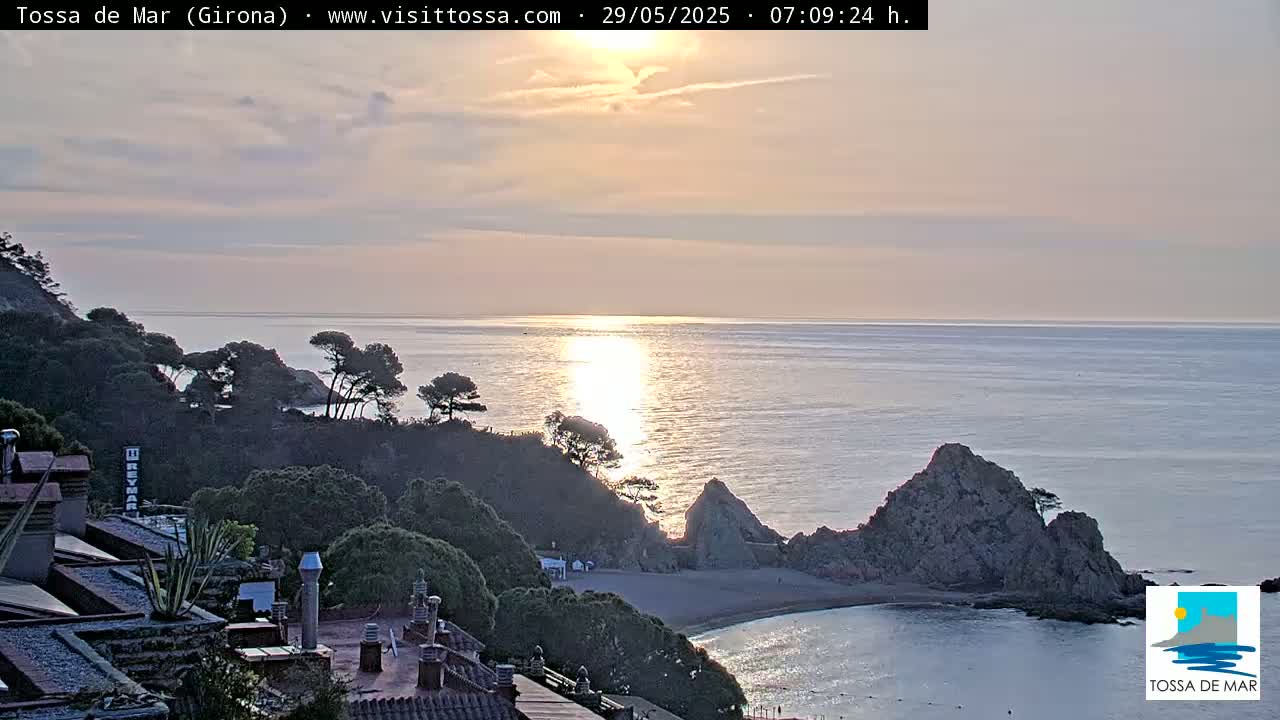 A sunrise over a calm sea, illuminating a rocky cove with a small beach nestled between green hills and buildings.