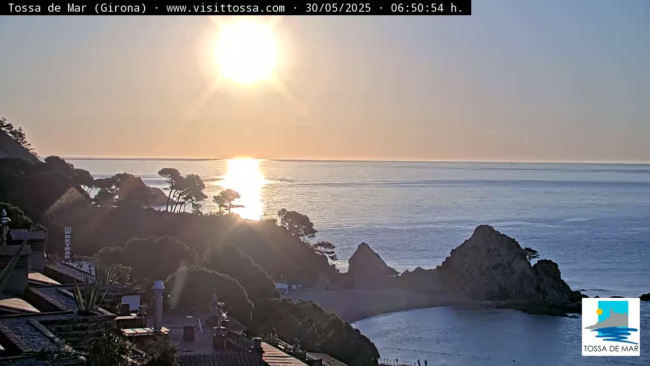 A coastal sunrise illuminates a rocky cove and calm ocean, with dark green vegetation covering the hillsides.