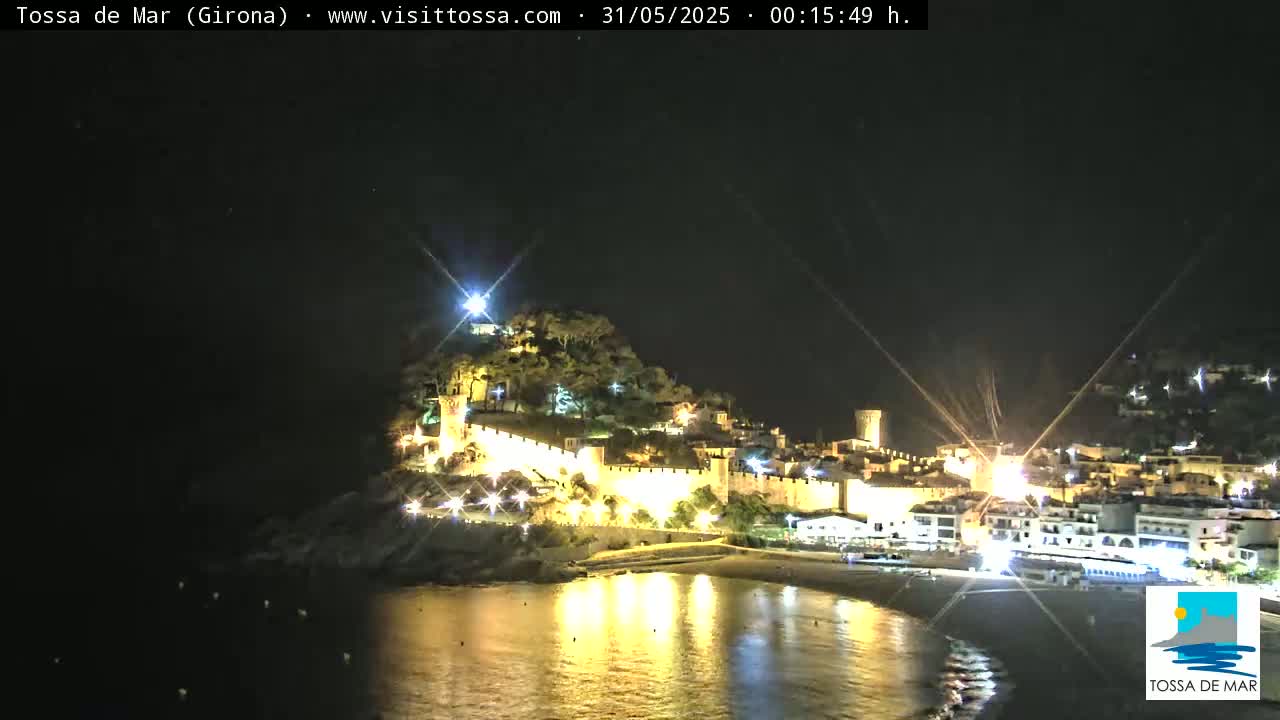 A hilltop town with a walled perimeter is brightly lit at night, its lights reflecting on a calm, dark body of water.