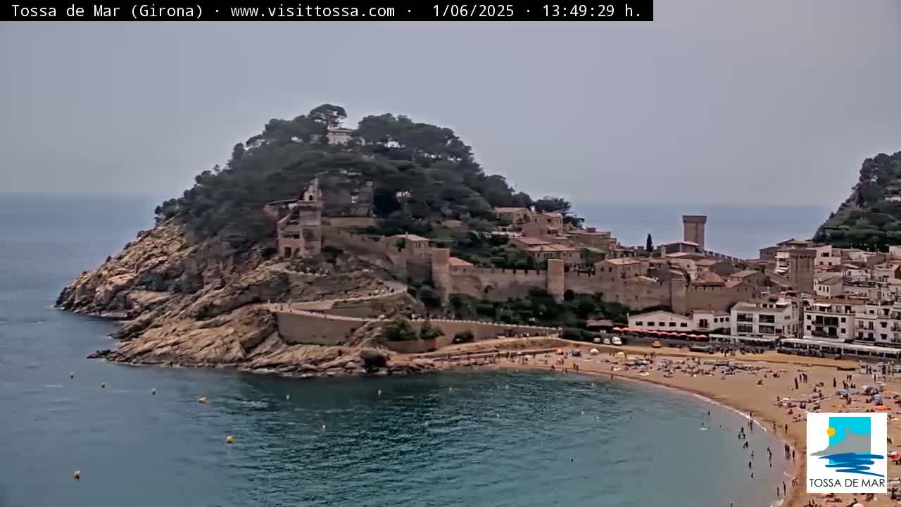 A coastal town with a medieval castle sits atop a hill overlooking a calm bay with a sandy beach, under an overcast sky.