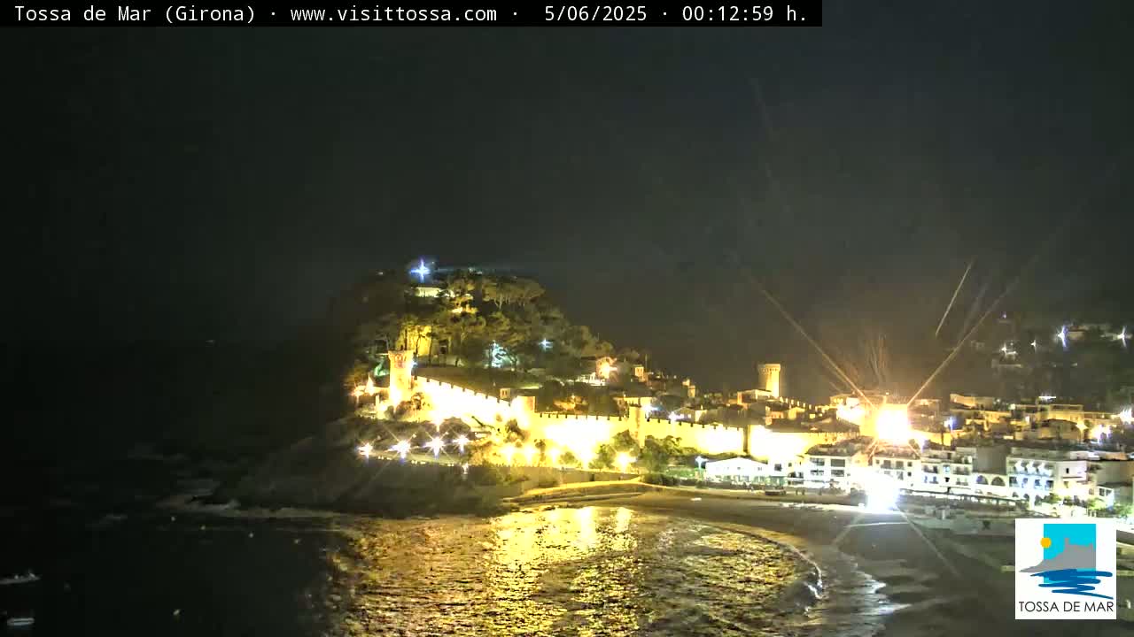 A nighttime coastal town with a walled fortress on a hill is illuminated, reflecting in the dark water below under a clear sky.