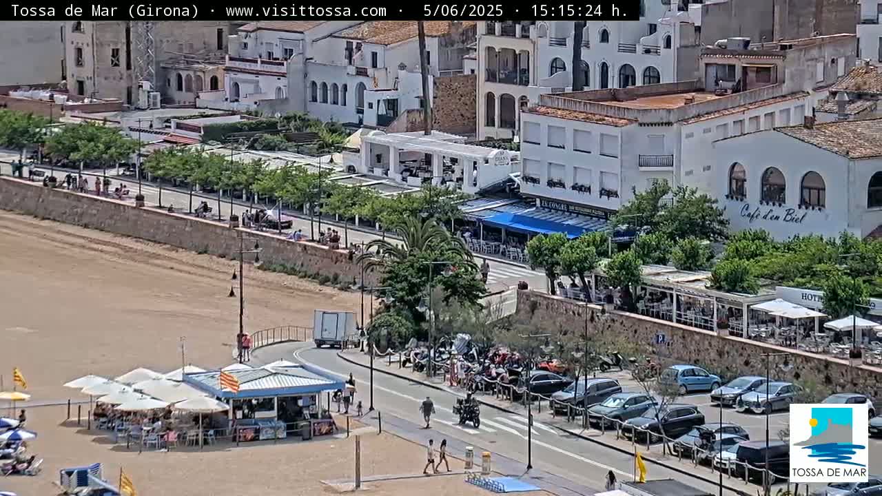 A sunny beach scene shows a sandy shore with beach umbrellas and chairs, a seaside promenade with restaurants and parked cars, and white buildings in the background.