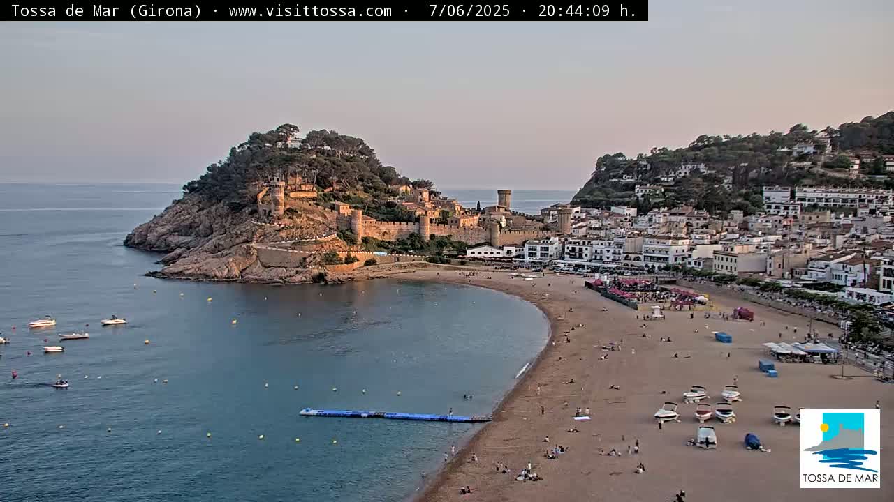 A coastal town with a medieval castle perched atop a cliff overlooks a sandy beach and calm bay under a clear evening sky.