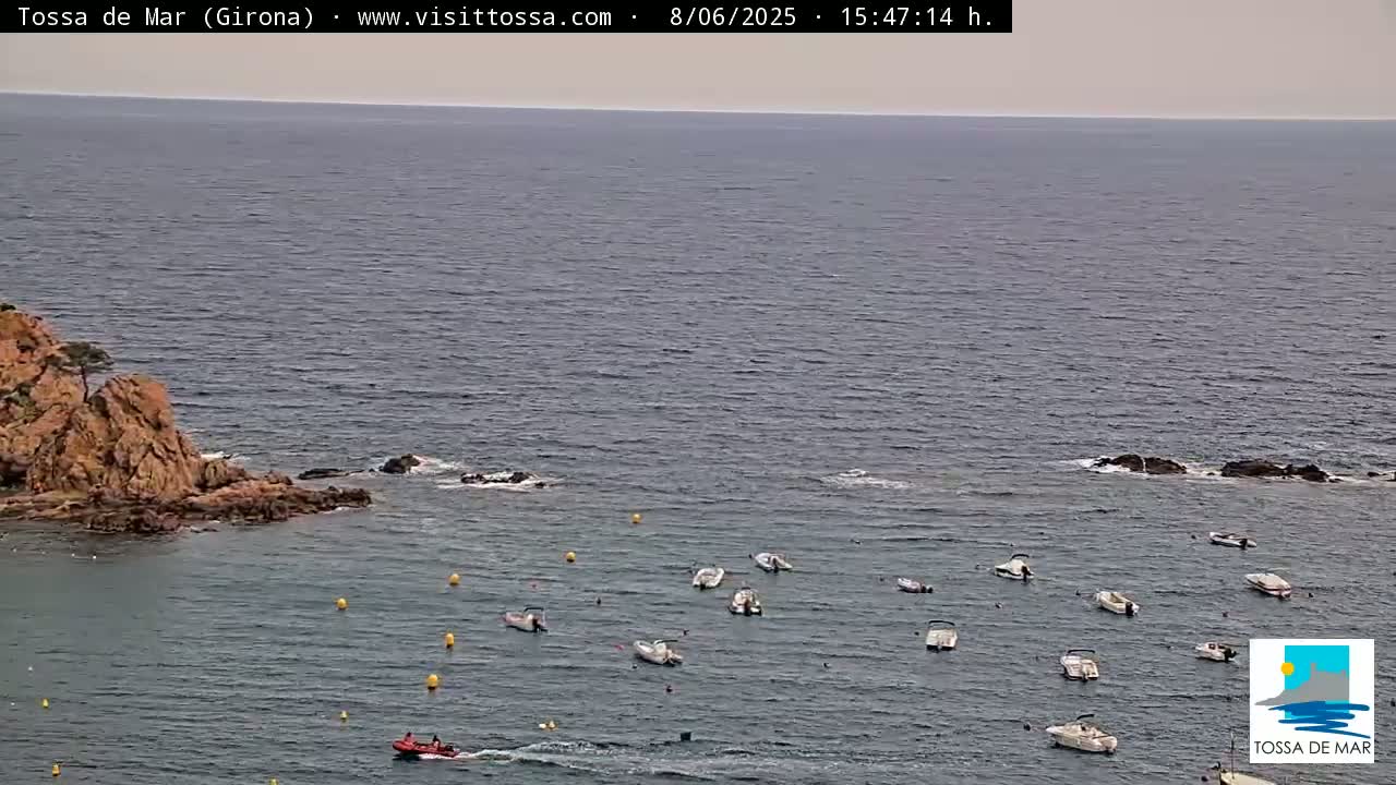 A rocky shoreline juts into a calm, gray sea where numerous small boats are anchored.