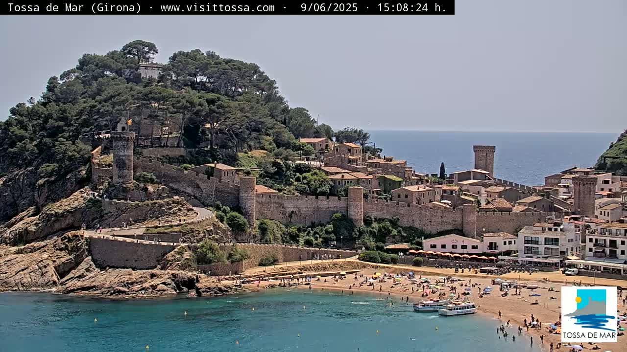 A walled medieval town sits on a rocky cliff overlooking a sandy beach and calm blue sea under a bright, sunny sky.