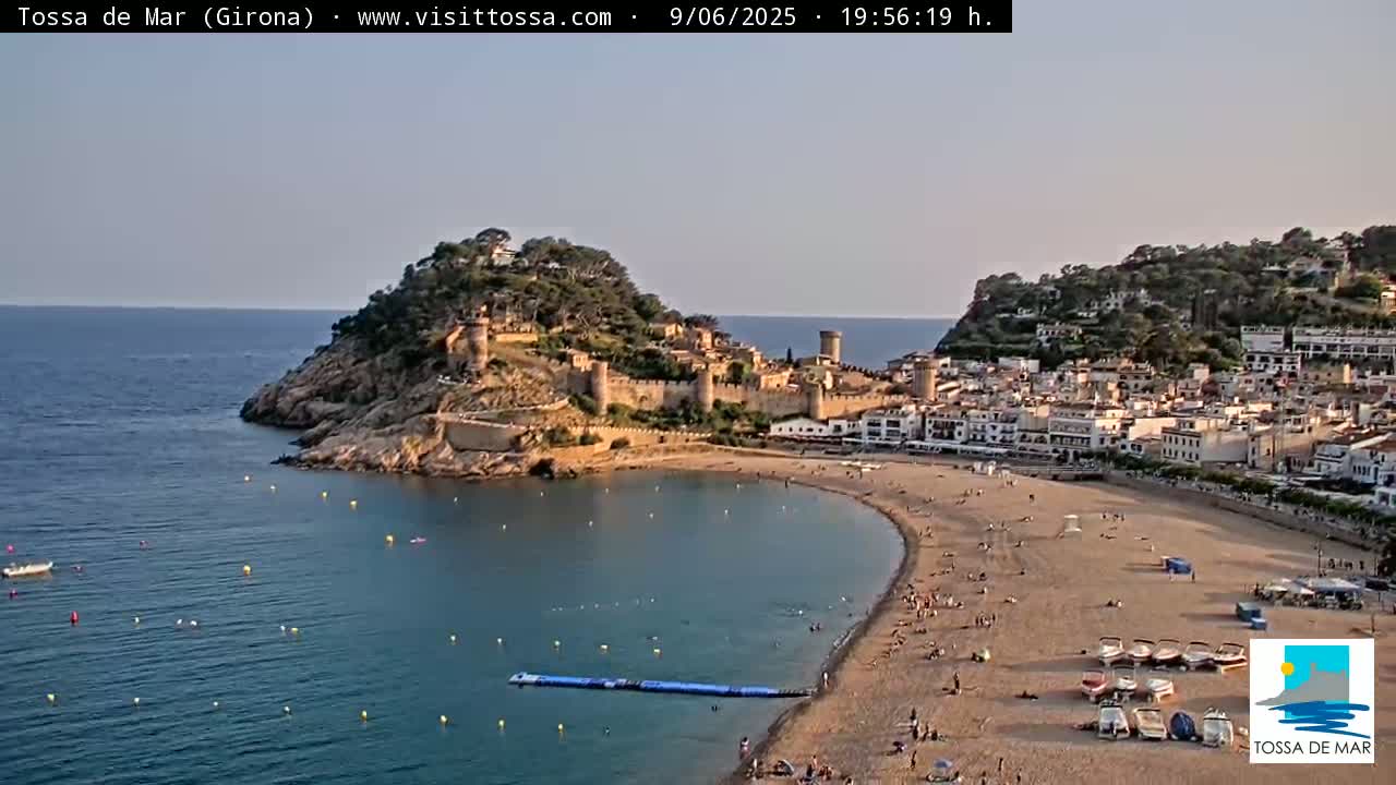 A coastal town with a medieval castle on a hill overlooking a sandy beach and calm sea under a clear, twilight sky.