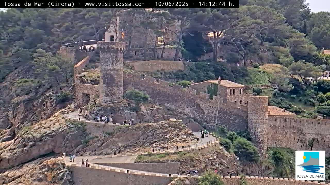 A stone fortress winds along a rocky hillside overlooking a lush, green landscape under a bright, sunny sky.
