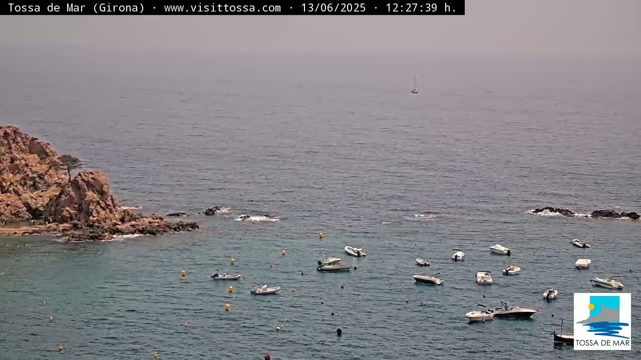 A hazy day shows a rocky cove with numerous small boats moored in calm, clear water, and a single sailboat further out at sea.