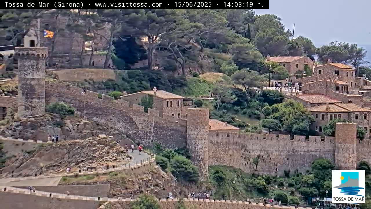 A stone castle wall surrounds a village of stone buildings nestled on a hillside on a partly cloudy day.
