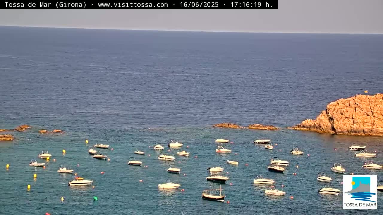 Numerous small boats are moored in a calm, clear body of water near a rocky outcrop on a sunny day.