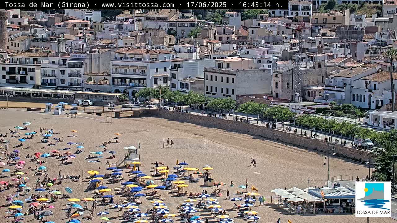 A sandy beach with numerous colorful umbrellas and sunbathers is situated below a town of closely packed buildings under a sunny sky.