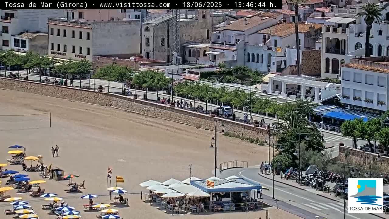 A sandy beach with beach umbrellas and a beachside restaurant is seen next to a town with buildings, palm trees, and people walking along a paved road, on a sunny day.