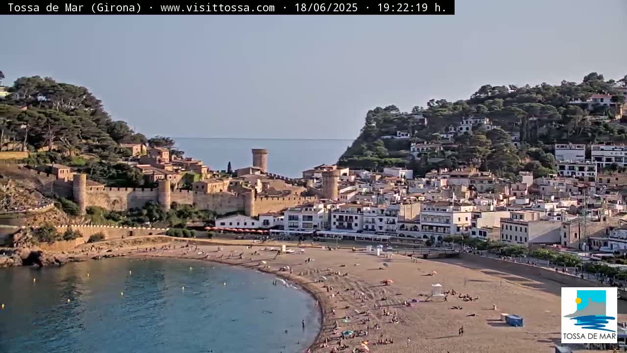 A coastal town with a medieval walled section and a sandy beach is visible under a clear sky.