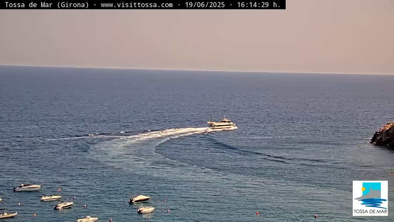A ferry leaves a wake as it travels across a calm, hazy sea where several small boats are anchored near a rocky shoreline.