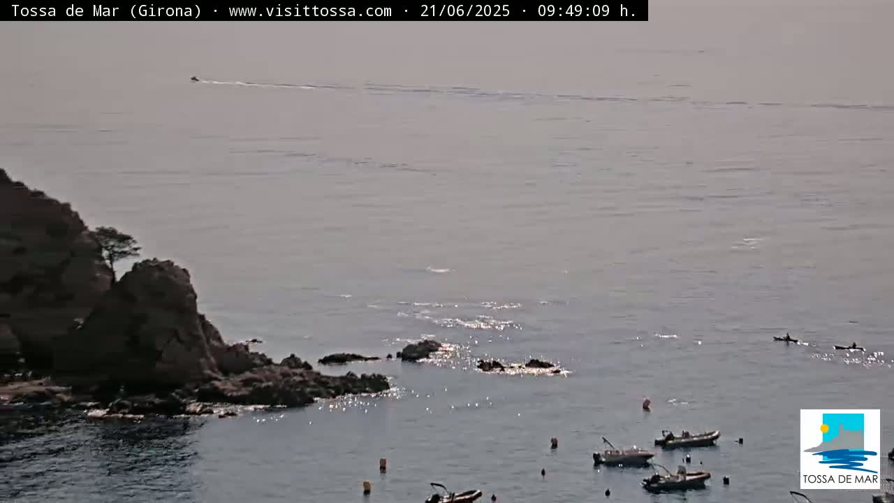 A rocky outcrop juts into a calm, slightly hazy sea where several small boats and kayaks are visible.